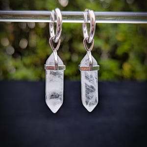 May include: A pair of silver hoop earrings with clear crystal point charms hanging from them. The earrings are against a blurred green background.
