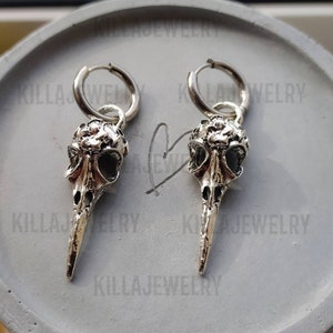 May include: A pair of silver hoop earrings with a bird skull charm hanging from each hoop. The skull charms have a detailed design and a long, pointed beak.