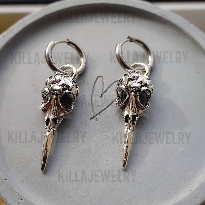 May include: A pair of silver hoop earrings with a bird skull charm hanging from each hoop. The skull charms have a detailed design and a long, pointed beak.