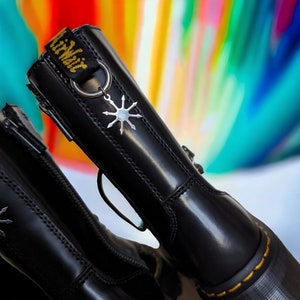 Op de afbeelding: Een paar zwarte leren laarzen met een rits aan de zijkant en een zilveren stervormige hanger. De laarzen hebben een gele stikseldetail rond de zool.