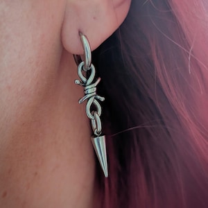 May include: A silver earring featuring a barbed wire design and a dangling, cone-shaped spike. The earring is attached to a silver hoop. The background shows pink hair and skin.