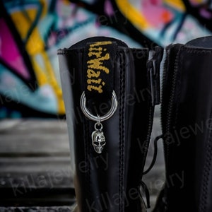 May include: Black leather boots with a silver skull charm attached to a ring on the zipper pull. The boots have a yellow and black Dr. Martens logo on the back.