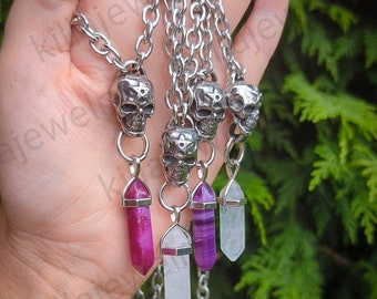 May include: Five silver skull necklaces with crystal pendants. The pendants are clear, purple, and pink. The necklaces are on a hand with painted nails.