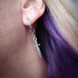 May include: A silver hoop earring with a dangling silver sword charm. The earring is worn in a person's ear.