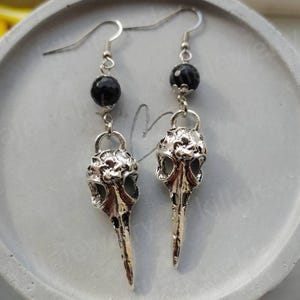 May include: A pair of silver bird skull earrings with black beads. The earrings are dangling and have a detailed skull design.