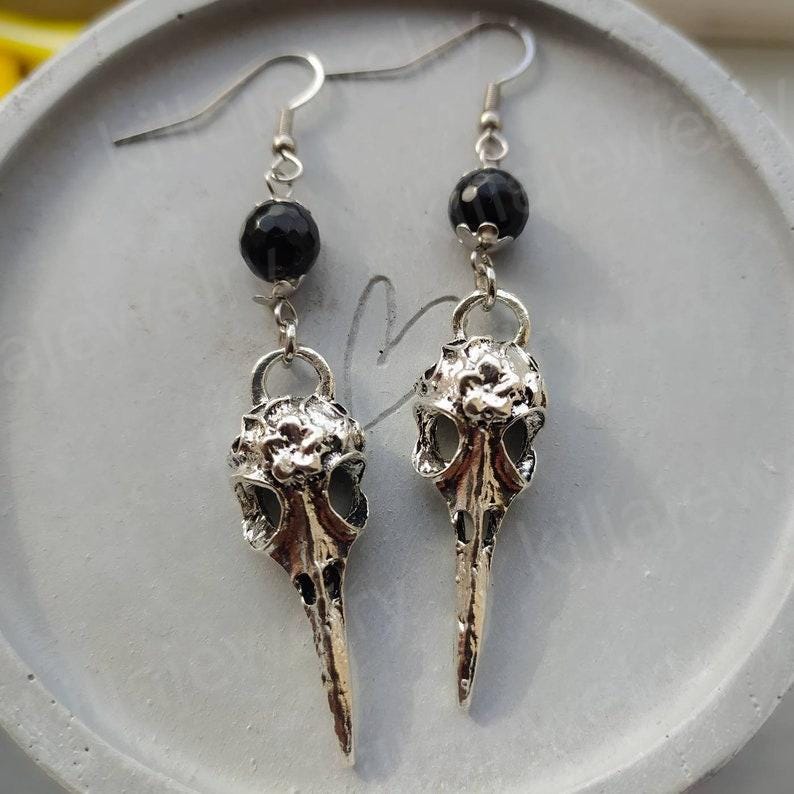 May include: A pair of silver bird skull earrings with black beads. The earrings are dangling and have a detailed skull design.