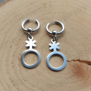 以下が含まれることがあります： ジェンダーシンボルが描かれたチャームがぶら下がった、銀色のフープイヤリングのペア。シンボルは、十字架が内側に描かれた円形です。