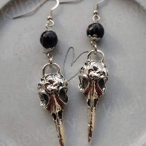 May include: Silver bird skull earrings with black beads. The earrings are dangling and have a detailed design.