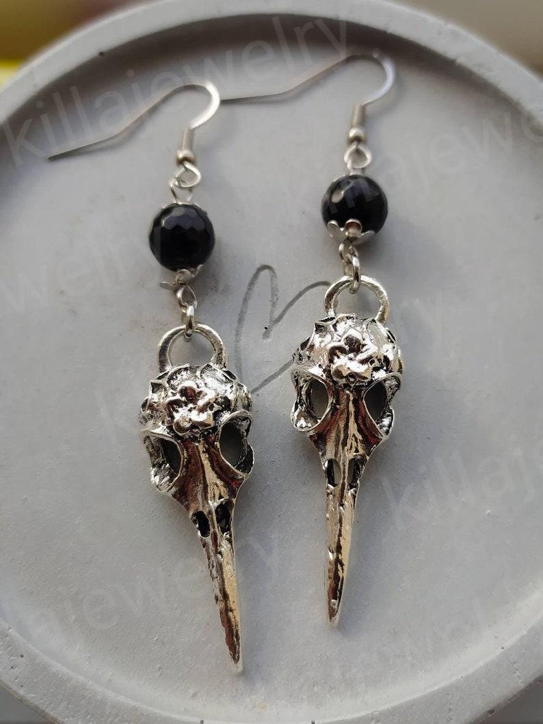 May include: Silver bird skull earrings with black beads. The earrings are dangling and have a detailed design.