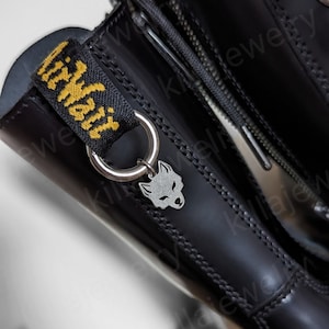 Peut inclure: Gros plan sur une botte en cuir noir avec un charm loup argenté suspendu à un anneau métallique. La botte a une fermeture éclair et une tirette en tissu noir et jaune avec l'inscription "Air Wair". La botte est brillante et présente des coutures visibles.