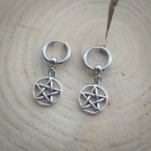 Peut inclure: Une paire de boucles d'oreilles cr&eacute;oles argent&eacute;es. Chaque boucle d'oreille est compos&eacute;e d'un cercle avec une petite boule en haut et d'un pendentif en forme de pentagramme. Les boucles d'oreilles sont pos&eacute;es sur une surface en bois brun clair.