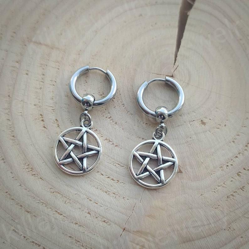 Peut inclure: Une paire de boucles d'oreilles cr&eacute;oles argent&eacute;es. Chaque boucle d'oreille est compos&eacute;e d'un cercle avec une petite boule en haut et d'un pendentif en forme de pentagramme. Les boucles d'oreilles sont pos&eacute;es sur une surface en bois brun clair.