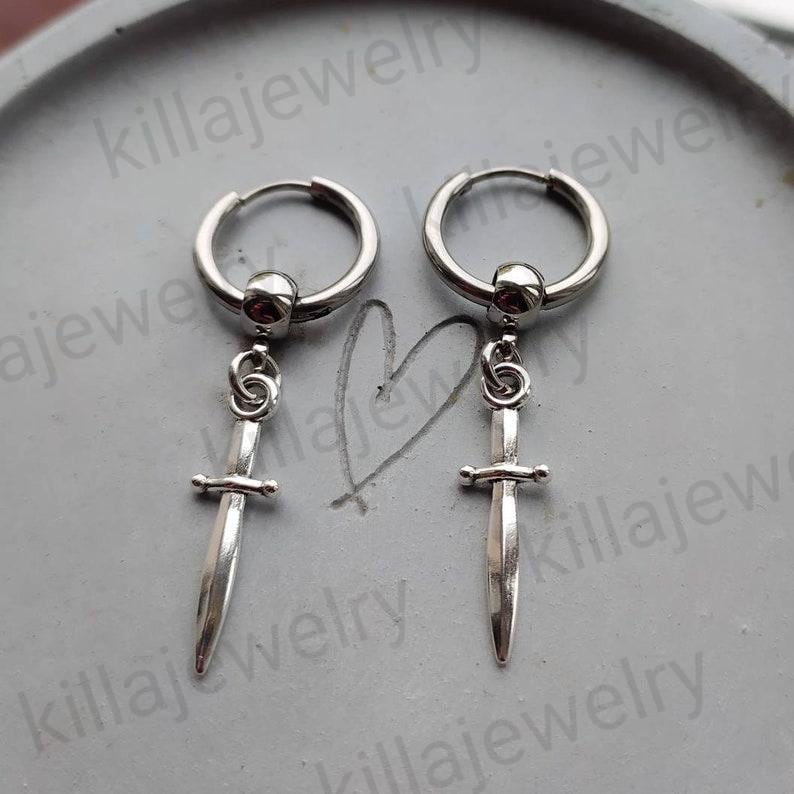 May include: A pair of silver hoop earrings with a dangling sword charm. The swords have a detailed design and are silver in color.