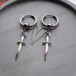 May include: A pair of silver hoop earrings with a dangling sword charm. The swords have a silver finish and a red gem at the top of the hoop.