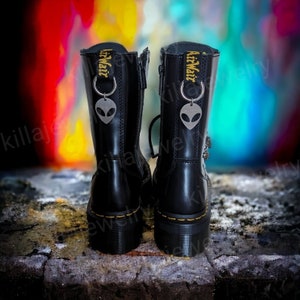 May include: Black leather boots with a platform sole and a zipper up the back. The boots have a yellow tag with the text "AirWair" on them and a silver alien charm attached to the tag.