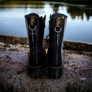 May include: Black leather boots with a zipper up the back and a silver charm with a crescent moon and a sword hanging from the top of the zipper. The boots have the text "AirWair" printed on the back of the boot.