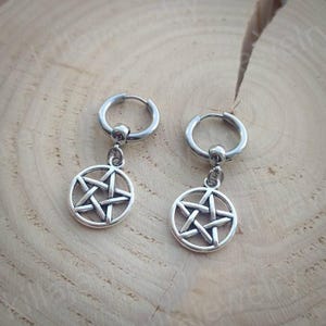 Peut inclure: Une paire de boucles d'oreilles cr&eacute;oles argent&eacute;es. Chaque boucle d'oreille est orn&eacute;e d'un pendentif circulaire avec un motif d'&eacute;toile &agrave; cinq branches. Les boucles d'oreilles sont pr&eacute;sent&eacute;es sur une surface en bois clair et textur&eacute;.