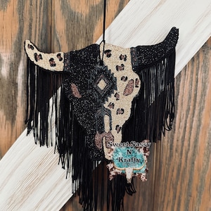 May include: Black and gold glitter cow skull with black fringe hanging from a black string. The skull has a geometric design in the center and a leopard print pattern on the side. The skull is labeled "Sweet Sassy N Krafty Est 2020".