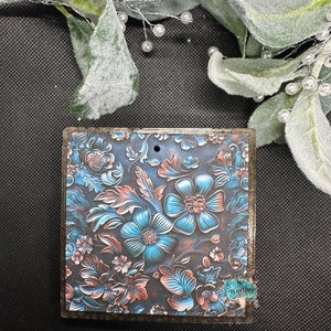 May include: A square wooden ornament with a blue and copper floral design. The ornament has a distressed wood finish and a small hole at the top for hanging.