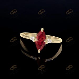 May include: A gold ring featuring a marquise-cut, deep red gemstone as the focal point. The ring is flanked by smaller, clear gemstones on each side. The band is a simple, polished gold. The ring is set against a black background.