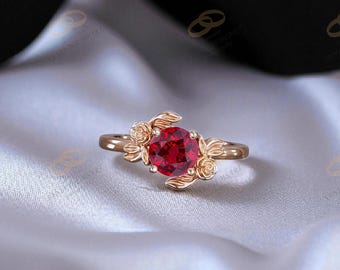 May include: Rose gold ring featuring a large, round, deep red gemstone. The ring's design incorporates floral elements, with rose-shaped accents on either side of the central stone. The band has a delicate, polished finish, and the overall aesthetic is elegant.