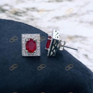 May include: A pair of silver stud earrings. Each earring features a rectangular frame of small, clear gemstones surrounding a large, oval-shaped, red gemstone. The earring posts are visible.