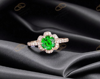 Puede incluir: Un anillo de oro rosa con una piedra preciosa verde vibrante en el centro, rodeada de piedras más pequeñas y brillantes. La banda del anillo también está adornada con piedras de acento, creando un diseño floral. El anillo se encuentra sobre un fondo negro.