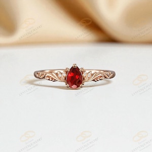 Puede incluir: Un anillo de oro rosa con una piedra preciosa roja oscura en forma de pera. El anillo tiene un diseño ornamentado con pequeñas piedras de acento claras y delicados detalles de filigrana en la banda. El anillo se muestra sobre un fondo neutro.