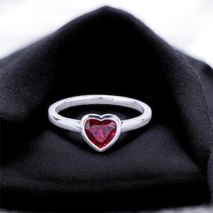 May include: A silver ring with a heart-shaped, deep red gemstone. The ring is set against a black background, highlighting the vibrant color of the stone. The band is thin and polished.