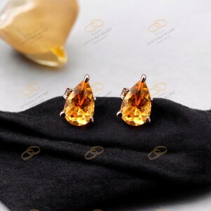 Könnte beinhalten: Ein Paar tropfenförmige Ohrringe mit einem leuchtend orange-gelben Edelstein. Die Ohrringe sind in Roségold gefasst und liegen auf einem schwarzen Stoff. Die Edelsteine sind facettiert, wodurch sie das Licht einfangen und funkeln.