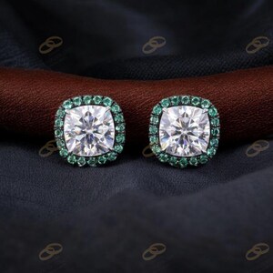 May include: A pair of square-cut stud earrings. Each earring has a large, clear, sparkling central stone encircled by a halo of small, green gemstones. The earrings are set against a dark background.