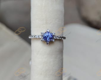 May include: A silver ring with a central, round, light blue gemstone, surrounded by smaller, clear stones in a starburst design. The band is also adorned with small, clear stones. The ring is displayed on a white, cylindrical surface.