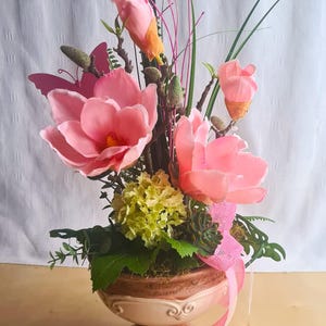 Könnte beinhalten: Dekorative Blumengesteck mit großen rosa Blüten, Knospen und Grün in einer Vase. Die Vase ist hellbraun mit cremefarbenem Oberteil und rosa Schleife. Ein rosa Schmetterling sorgt für einen dekorativen Akzent.