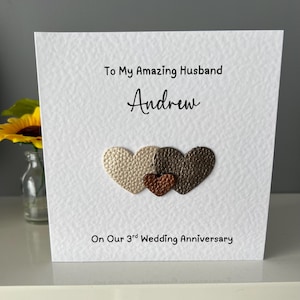 May include: A white textured anniversary card with the text "To My Amazing Husband Andrew" and "On Our 3rd Wedding Anniversary." The card features three heart-shaped embellishments in gold, brown, and copper colours.