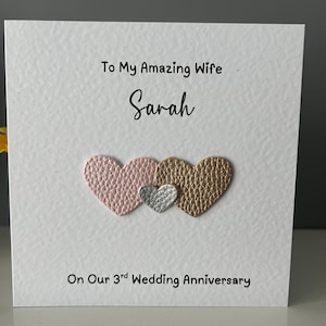 May include: A white card with the text "To My Amazing Wife Sarah" and "On Our 3rd Wedding Anniversary." The card features three heart-shaped embellishments in pink, silver, and gold.
