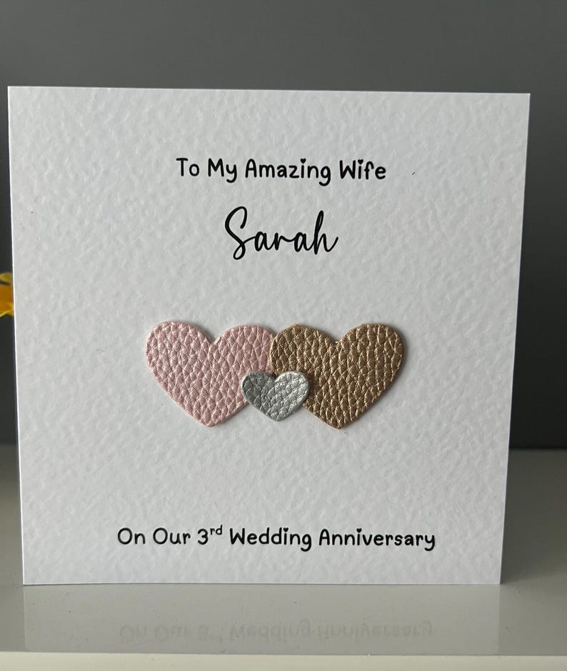 May include: A white card with the text "To My Amazing Wife Sarah" and "On Our 3rd Wedding Anniversary." The card features three heart-shaped embellishments in pink, silver, and gold.