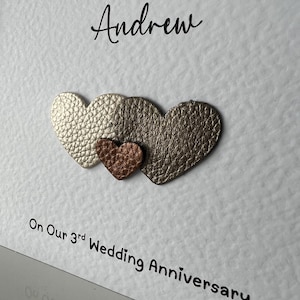 May include: A white card with the text "To My Amazing Husband Andrew" and "On Our 3rd Wedding Anniversary". Three heart-shaped embellishments in gold and copper tones are affixed to the card.