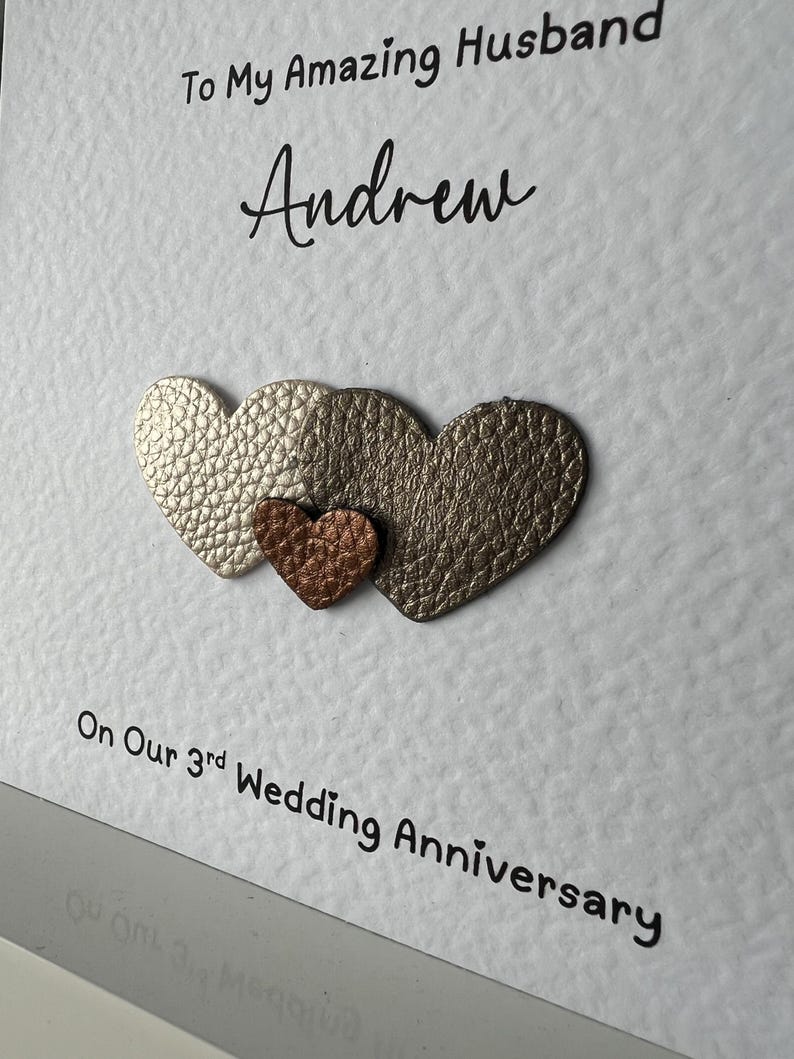 May include: A white card with the text "To My Amazing Husband Andrew" and "On Our 3rd Wedding Anniversary". Three heart-shaped embellishments in gold and copper tones are affixed to the card.