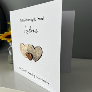 May include: A white greeting card with the text "To My Amazing Husband Andrew" and "On Our 3rd Wedding Anniversary." The card features three textured heart shapes in gold and bronze tones.