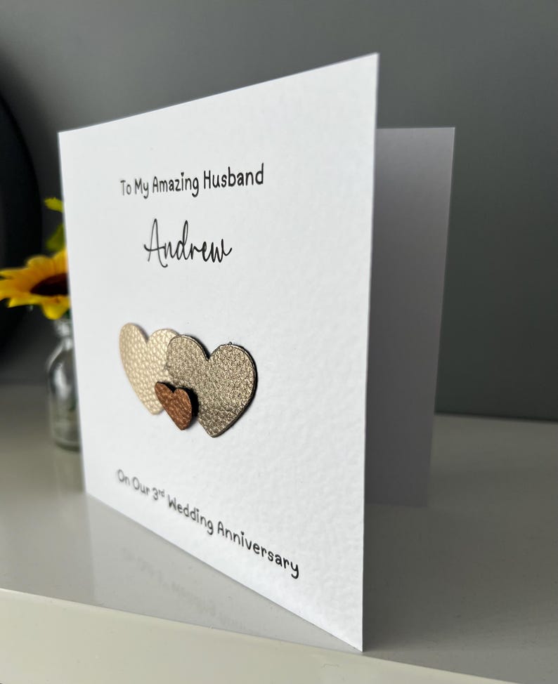 May include: A white greeting card with the text "To My Amazing Husband Andrew" and "On Our 3rd Wedding Anniversary." The card features three textured heart shapes in gold and bronze tones.