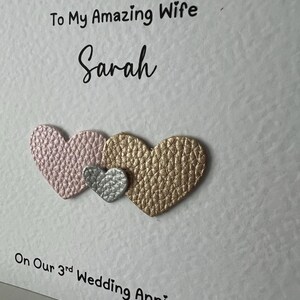 May include: A white card with the text "To My Amazing Wife Sarah" and "On Our 3rd Wedding Anniversary." Three textured hearts in pink, silver, and gold are below the text. A romantic greeting card.