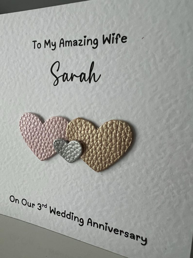 May include: A white card with the text "To My Amazing Wife Sarah" and "On Our 3rd Wedding Anniversary." Three textured hearts in pink, silver, and gold are below the text. A romantic greeting card.