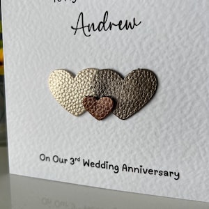 May include: A white greeting card with the text "To My Amazing Husband Andrew" and "On Our 3rd Wedding Anniversary." The card features three heart-shaped embellishments in gold, bronze, and copper colors.