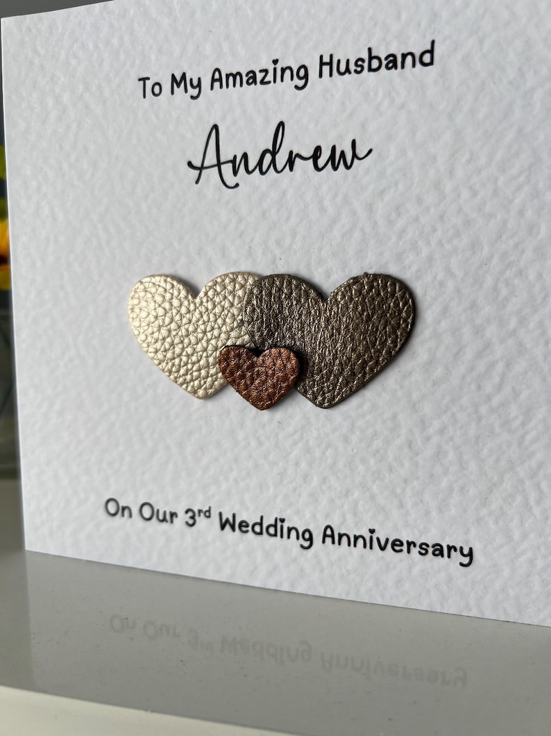 May include: A white greeting card with the text "To My Amazing Husband Andrew" and "On Our 3rd Wedding Anniversary." The card features three heart-shaped embellishments in gold, bronze, and copper colors.