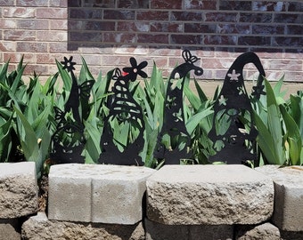 May include: Four black metal garden gnomes with whimsical designs stand in a row in front of a brick wall. The gnomes are surrounded by green plants.