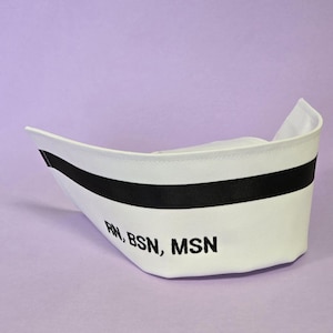 May include: A white nurse's cap with a black band and the embroidered letters "RN, BSN, MSN". The cap is set against a light purple background. The cap is a traditional style.