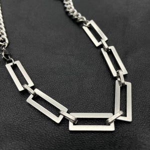 Puede incluir: Un collar de cadena de plata con eslabones rectangulares. La cadena está hecha de metal y tiene un acabado pulido.