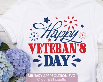 May include: A white short-sleeved t-shirt featuring a graphic design in red and blue. The text reads Happy VETERAN'S DAY in a mix of script and bold fonts, accented by small red and blue stars and firework-like bursts above and below the text.