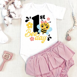 May include: White baby onesie with a black number one and the text "1st Bee-day" in yellow. A cartoon bee with a pink bow is next to the number one. The onesie is on a white background with a pink ruffled diaper cover.