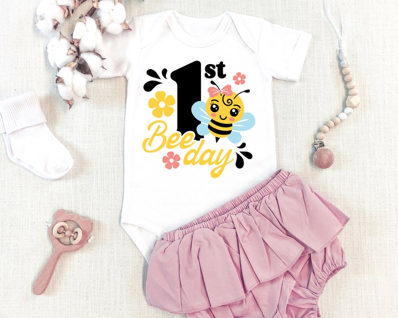 May include: White baby onesie with a black number one and the text "1st Bee-day" in yellow. A cartoon bee with a pink bow is next to the number one. The onesie is on a white background with a pink ruffled diaper cover.